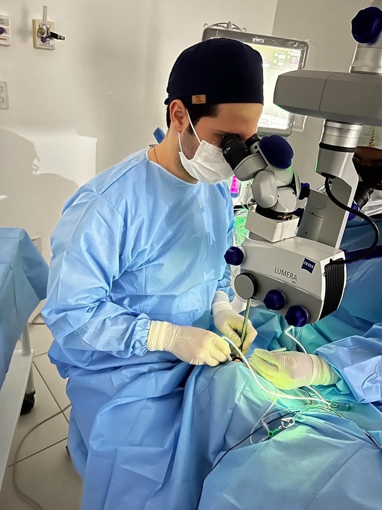 O Dr. Henrique Sampaio, oftalmologista, está realizando uma cirurgia de catarata. Ele está vestido com equipamento cirúrgico estéril, incluindo uma máscara e luvas. Ele está focado no procedimento, usando um microscópio cirúrgico para visualizar o olho do paciente. A imagem transmite a precisão e o cuidado necessários neste tipo de procedimento médico.