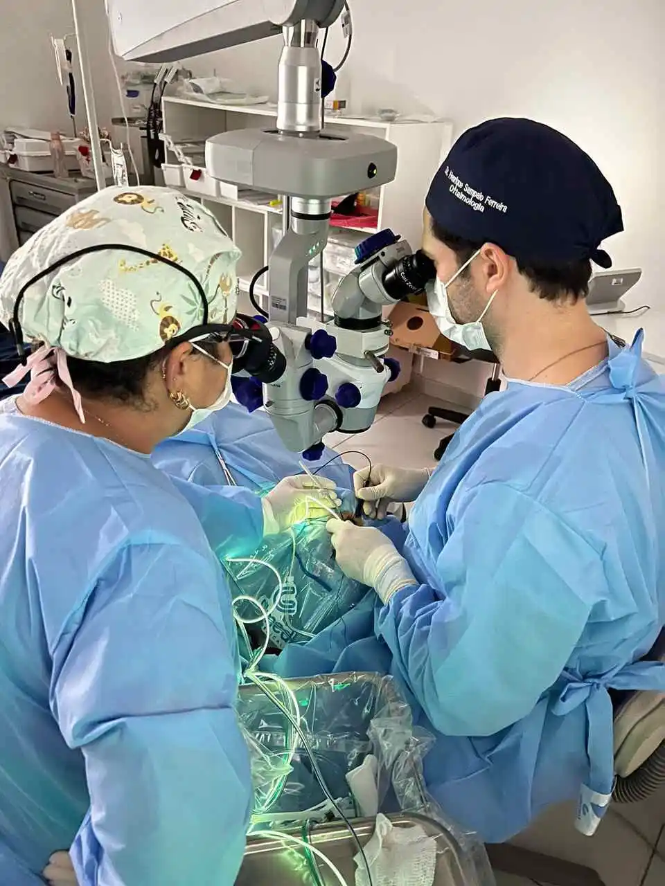 A imagem mostra dois indivíduos em trajes médicos realizando um procedimento cirúrgico. Eles estão cercados por equipamentos médicos, incluindo um microscópio. A sala é limpa e bem iluminada. Não é possível confirmar se o Dr. Henrique Sampaio está na imagem ou se a técnica de Vitrectomia está sendo usada.