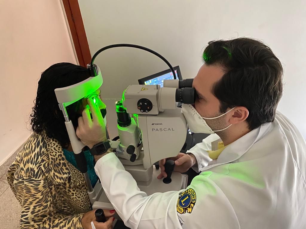 A imagem mostra um profissional médico e um paciente durante um exame ou procedimento de tratamento ocular. O profissional médico está usando um equipamento especializado com o nome da marca “PASCAL” visível nele. O paciente está sentado e tem a cabeça posicionada no equipamento, que emite uma luz verde. Ambos os indivíduos estão dentro de um ambiente, conforme sugerido pela parede e pelo chão visíveis ao fundo. Há um emblema ou logotipo na manga do jaleco branco do profissional médico, mas não está claro o suficiente para identificar.